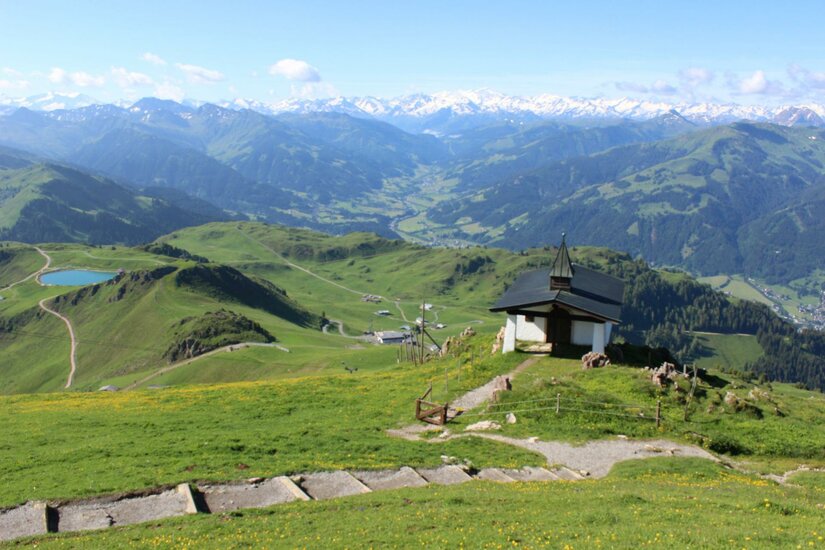Kapelle am Kitzbüheler Horn Kleine Kapelle auf einem grünen Berggipfel mit Blick auf ein weites Tal und schneebedeckte Alpen im Hintergrund.